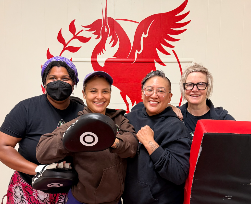6-Week Self-Defense Fundamentals: Love in Action Teaching Team - Photo of four smiling people standing in front of a wall with an illustration of a red crane, holding pads and other martial arts equipment